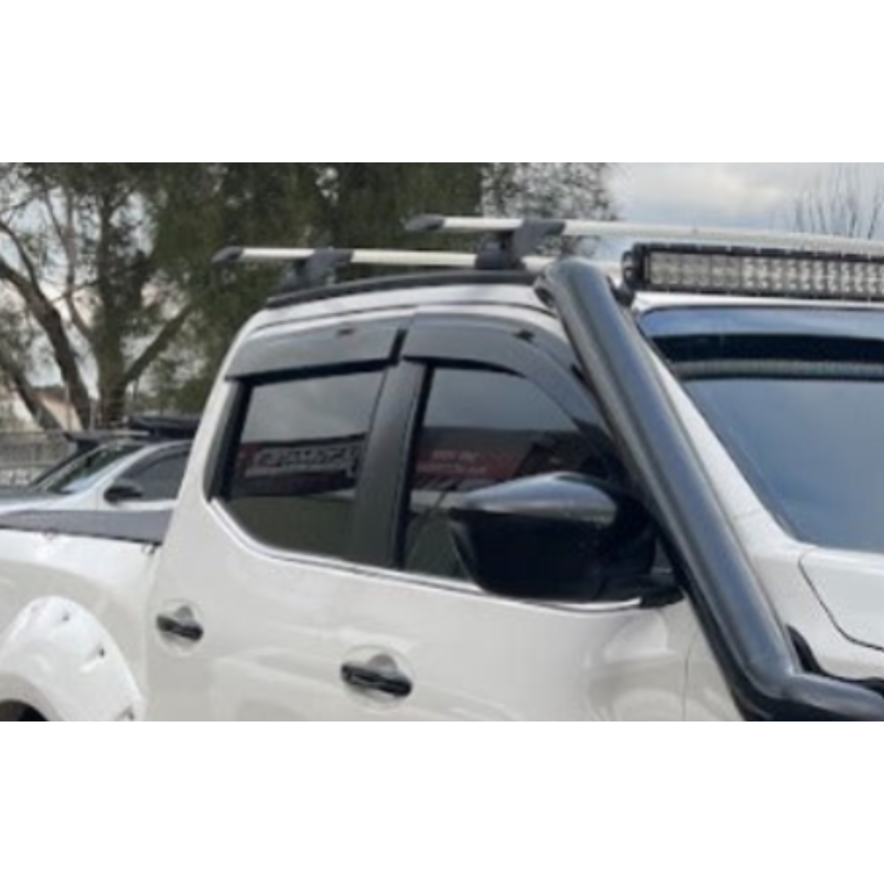 A close-up image of the front section of a white pickup truck, modified with several off-road accessories. Visible accessories include a large black air intake snorkel running up the side of the windshield, black plastic window visors/rain guards, a roof-mounted LED light bar, and silver roof rack cross bars. The truck also has black fender flares and dark tinted windows.