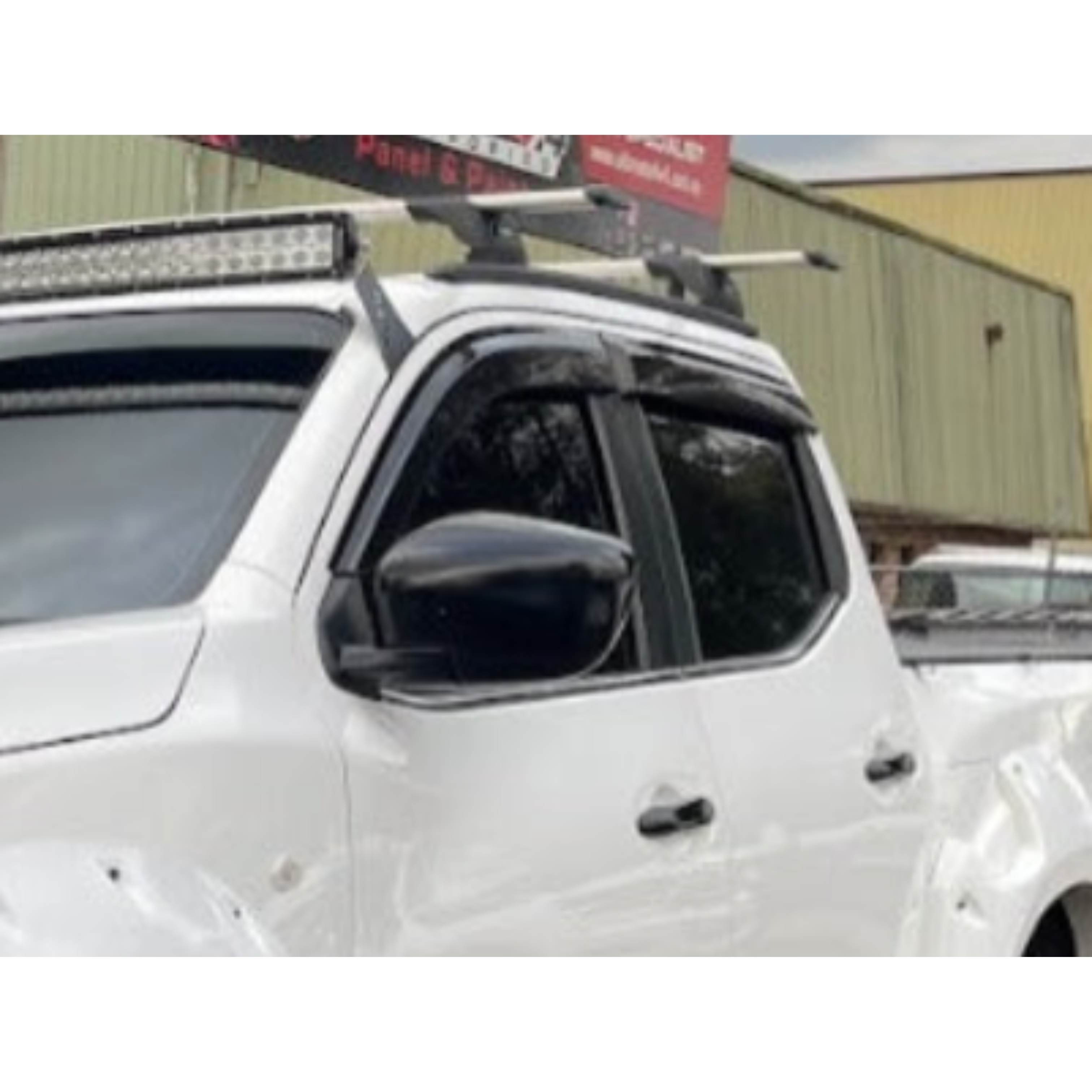 A close-up photograph of the cab of a white pickup truck. The truck is accessorized with a large LED light bar mounted above the windshield, silver and black roof rack cross bars, dark plastic window visors/rain guards on the side windows, and a black side mirror. A red and white sign is visible partially cropped at the top of the image.