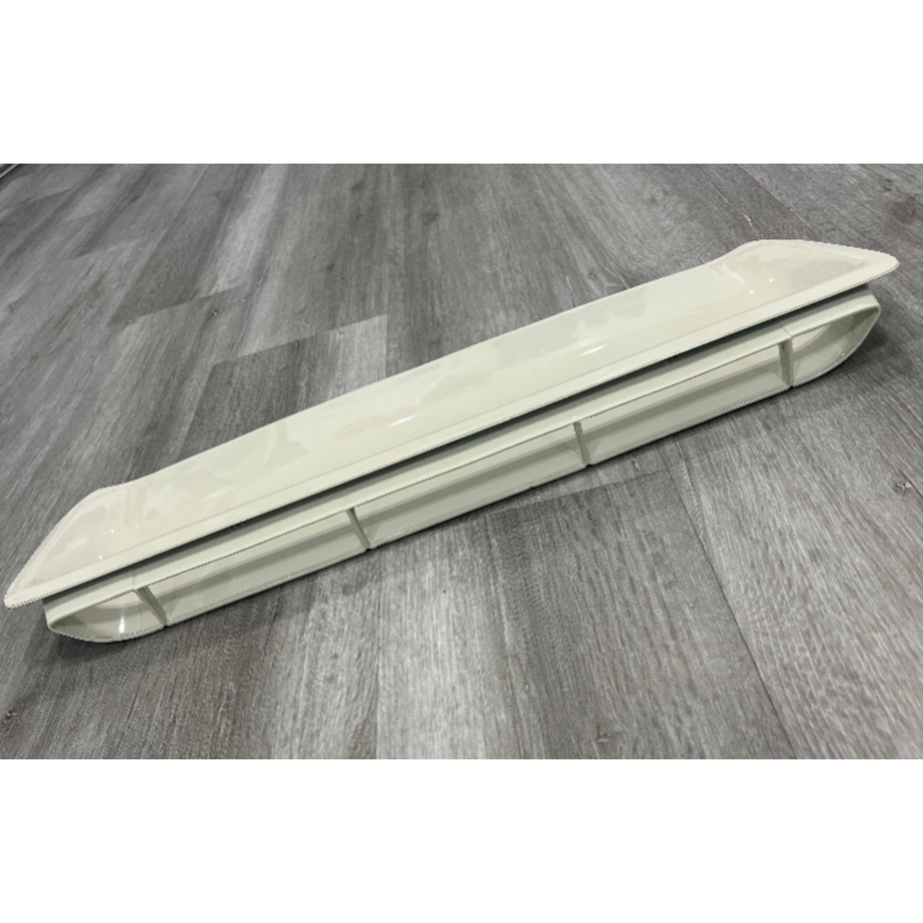 A long, beige or off-white plastic cover or housing, possibly for a light fixture or vent, is lying on a gray wood-look plank floor. The object is curved and elongated, with some internal or structural divisions visible along its side.