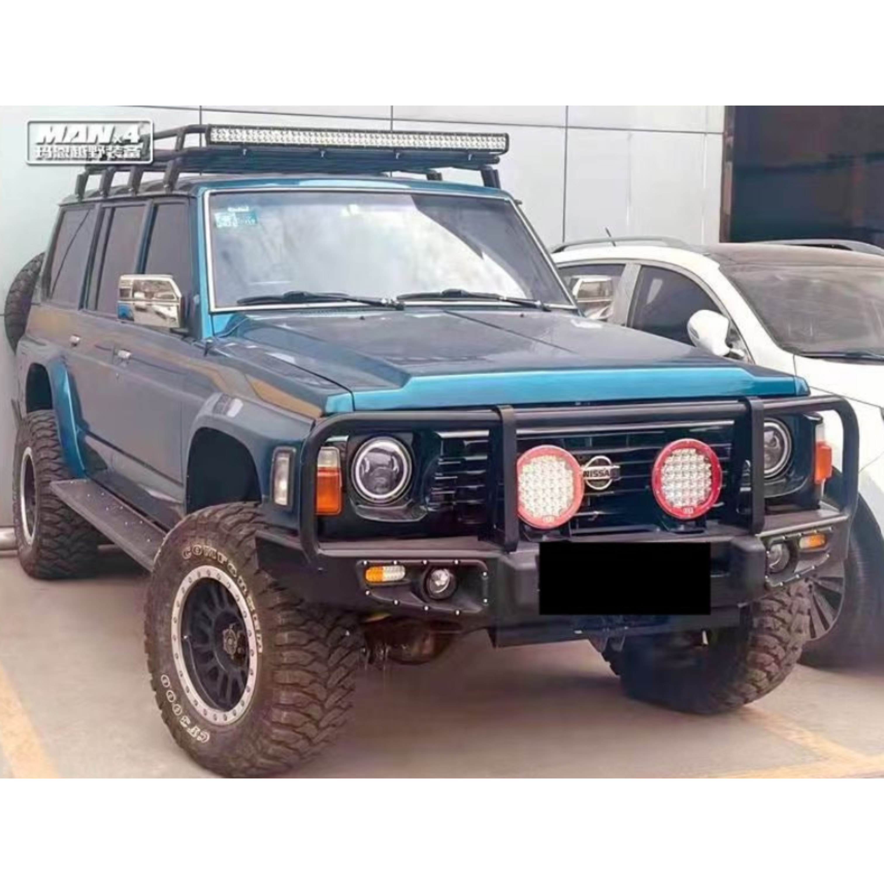 A highly modified, dark teal or blue-green Nissan Patrol Y60/GQ series SUV parked in a lot, showcasing numerous off-road accessories. Modifications include:

A heavy-duty steel bull bar (front bumper replacement) with three protective hoops.

Two large circular LED auxiliary driving lights with red trim mounted on the bull bar.

Aftermarket LED projector headlights.

A full-length basket roof rack with a large LED light bar mounted on the front.
