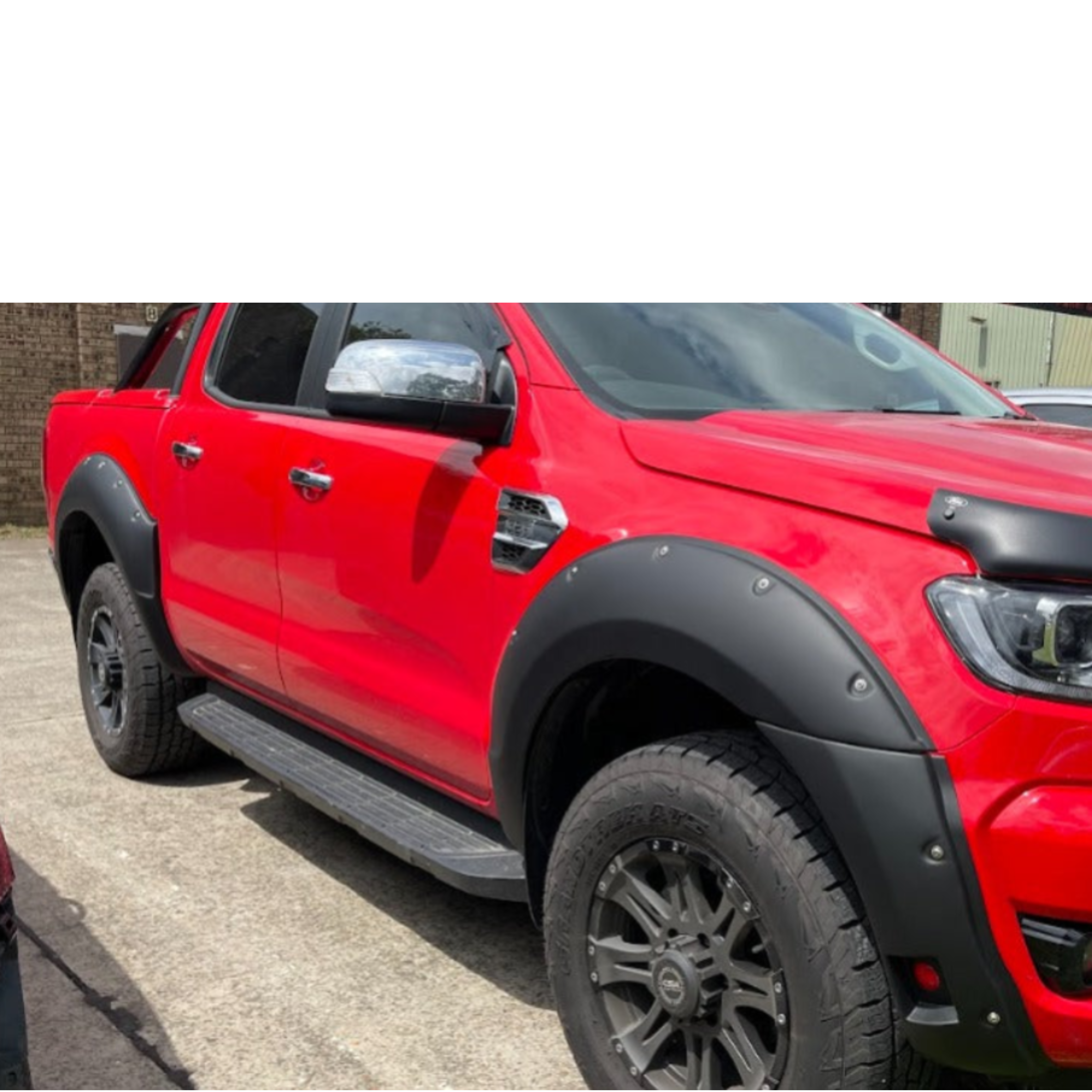 A side-angle view of a bright red Ford Ranger pickup truck modified with off-road accessories. The truck features matte black bolt-on style fender flares over all visible wheels and a wide, black textured side step/running board. It has black multi-spoke wheels with all-terrain tires and chrome side mirrors. A black hood protector is visible near the headlight.