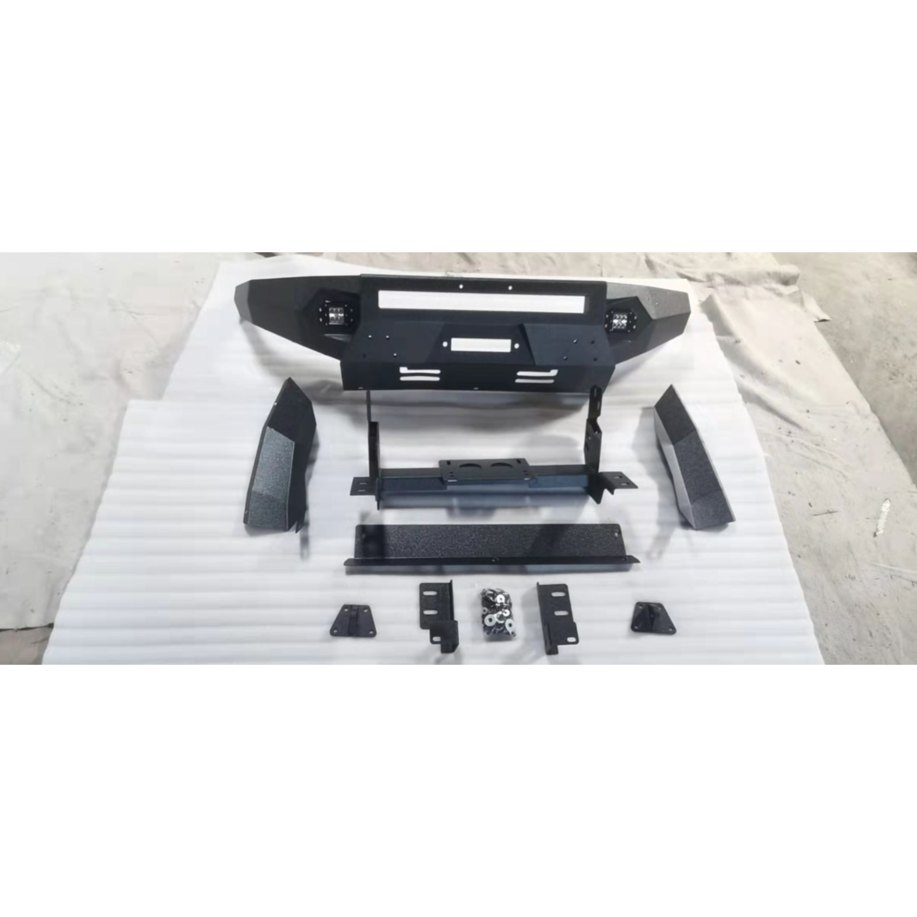 A disassembled, heavy-duty, matte black steel front bumper kit for an off-road vehicle is laid out on a white cloth surface. The main angular bumper section is at the top, featuring cutouts for a winch fairlead, a large central light bar opening, and two smaller recessed LED pod lights. The rest of the kit includes two textured, angled bumper end caps/side filler pieces, a central mounting bracket/cradle, a textured lower skid plate/valance piece, and several small mounting brackets and a bag of hardware.