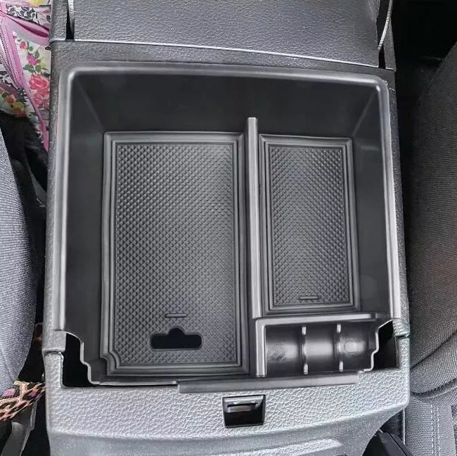 A plastic, black storage tray organizer placed inside a car's center console armrest compartment. The tray has two main sections with textured, diamond-patterned bottoms.
