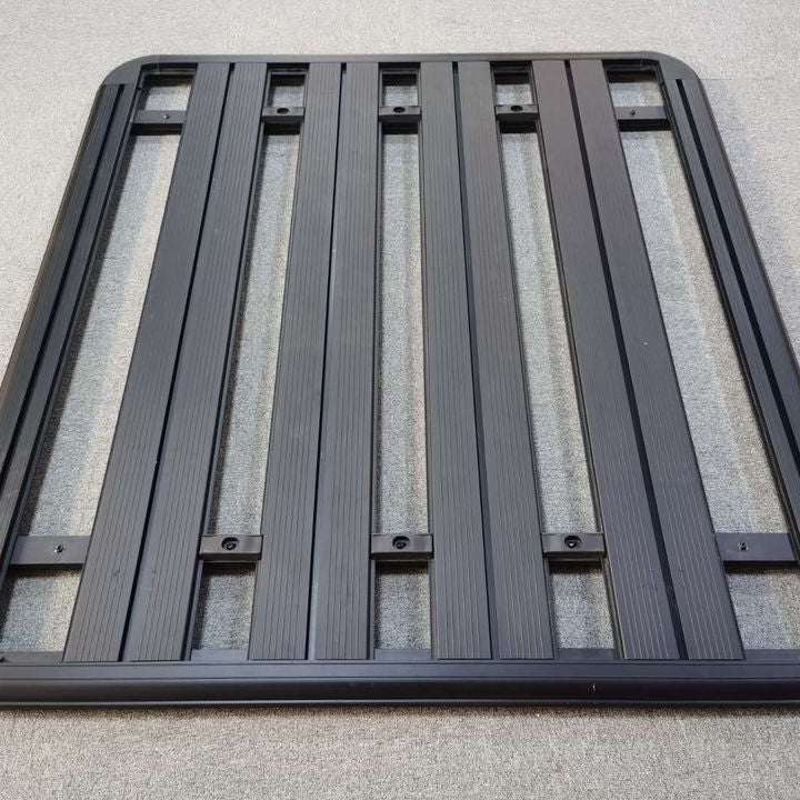 A rectangular, black, flat roof rack platform designed for a vehicle. The platform is constructed with multiple parallel, ribbed aluminum slats running lengthwise and is framed by a solid border. There are several recessed slots and mounting points visible along and between the slats, indicating a modular design for attaching accessories.