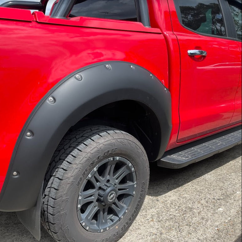 A close-up side view focusing on the rear wheel arch of a bright red pickup truck. The wheel arch is fitted with a wide, matte black bolt-on style fender flare with visible silver rivets. The truck has a black multi-spoke wheel and an all-terrain tire (Dunlop Grandtrek). A portion of a black side step/running board is also visible.