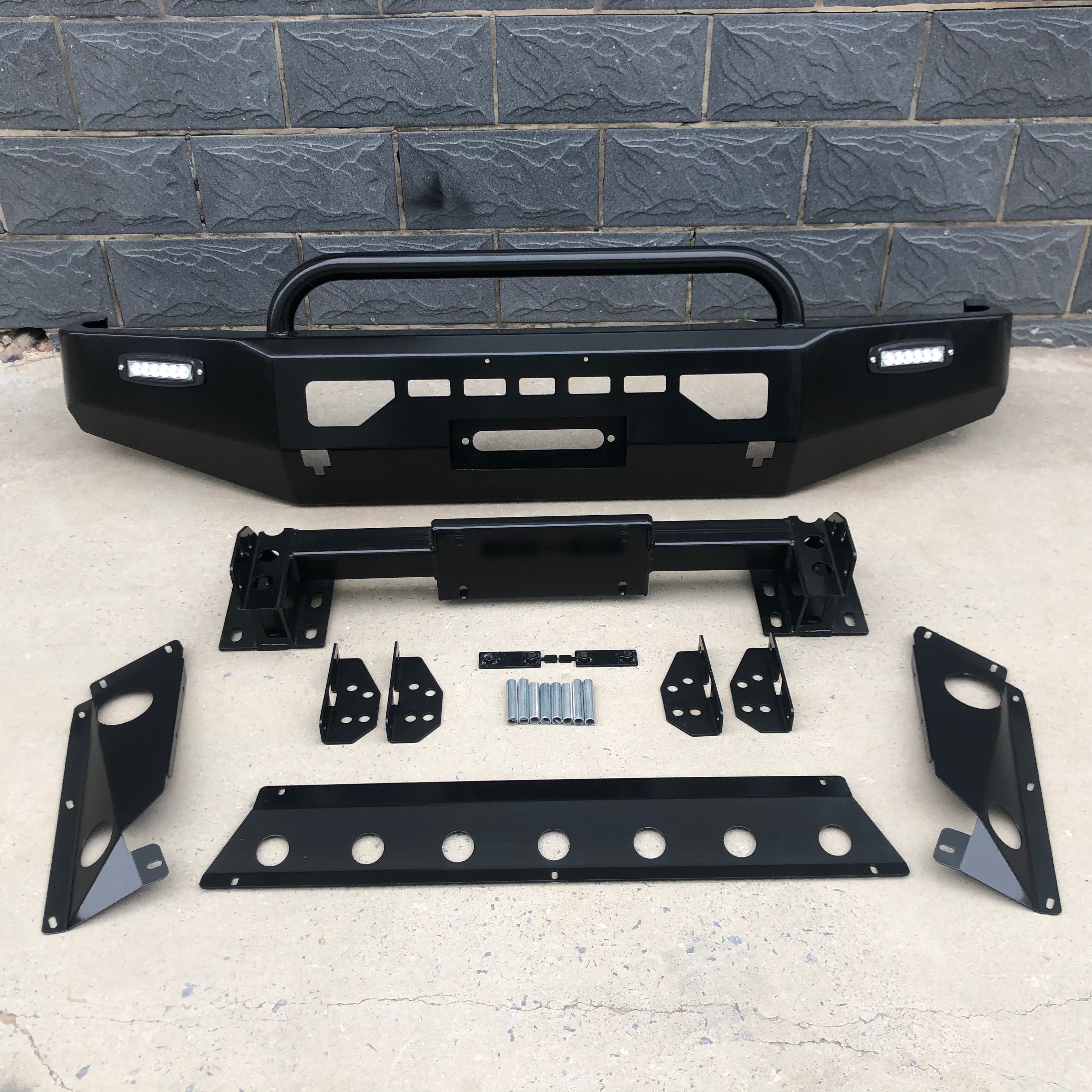 A complete black, heavy-duty aftermarket front bumper kit for a vehicle, displayed on a concrete floor in front of a dark gray cinder block wall. The main bumper assembly includes a large, integrated black tubular grille guard hoop and horizontal LED light bars mounted on the left and right outer edges. It also features a cutout for a winch fairlead. Below the main bumper are various smaller components, including a separate winch plate.