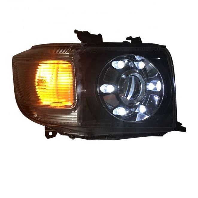A product photograph of a single aftermarket LED headlight assembly designed for a Toyota Land Cruiser 70 Series (or similar rectangular housing).

The housing is black and the cover is clear plastic.

The assembly features a rectangular amber turn signal (illuminated) on the left side.

The main headlight section on the right is round and includes a central projector lens surrounded by eight individual, bright white LED lights forming a ring or halo.

The unit is isolated on a white background.