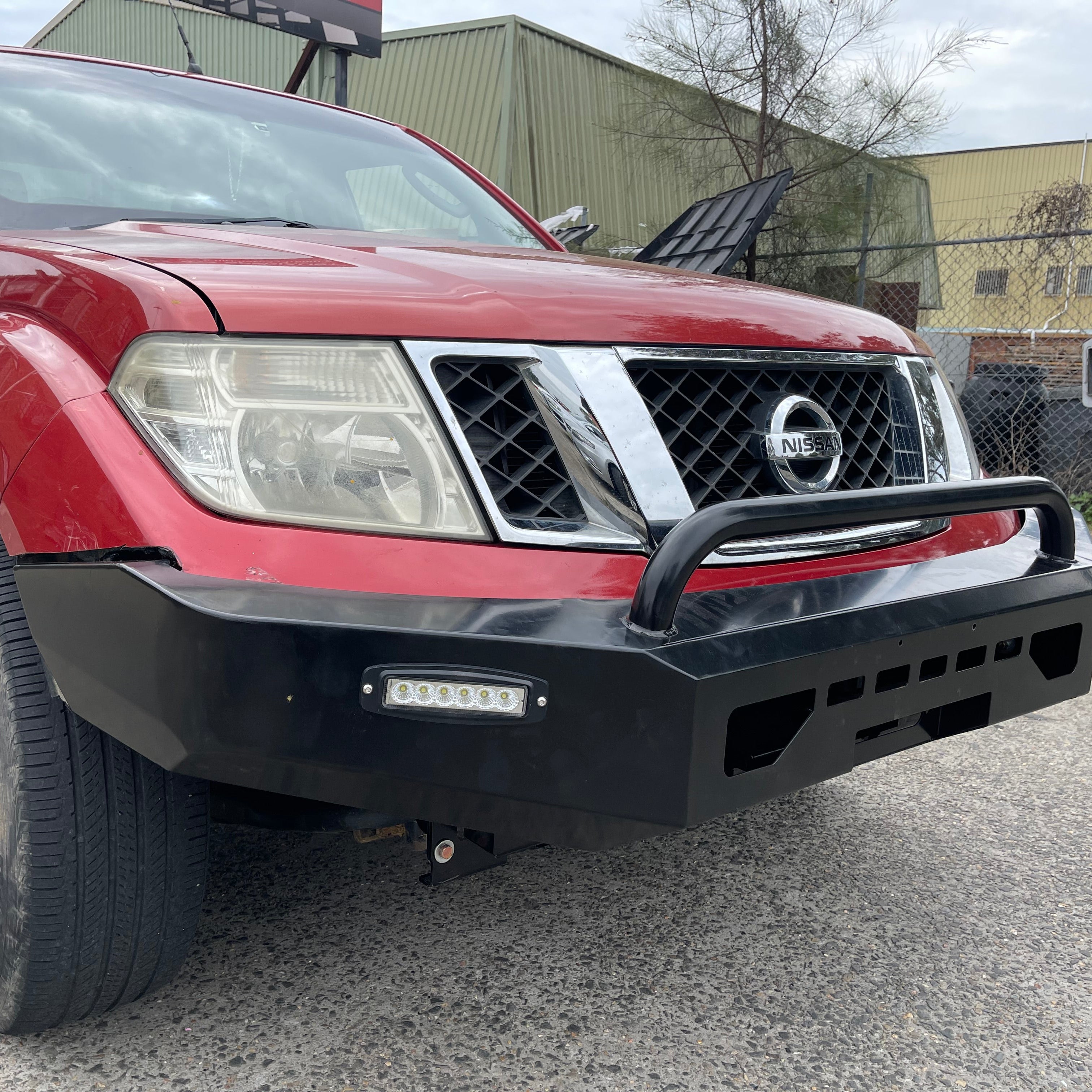 A close-up, low-angle view of the front end of a red Nissan Navara or Frontier (D40 generation) pickup truck. The truck is fitted with a heavy-duty, black aftermarket steel bull bar/bumper. The bumper has an angular design, features a single thick black tubular hoop protecting the grille, and an integrated horizontal LED light bar on the driver's side. The background is a slightly blurred industrial or outdoor area with a chain-link fence and a large metal building.
