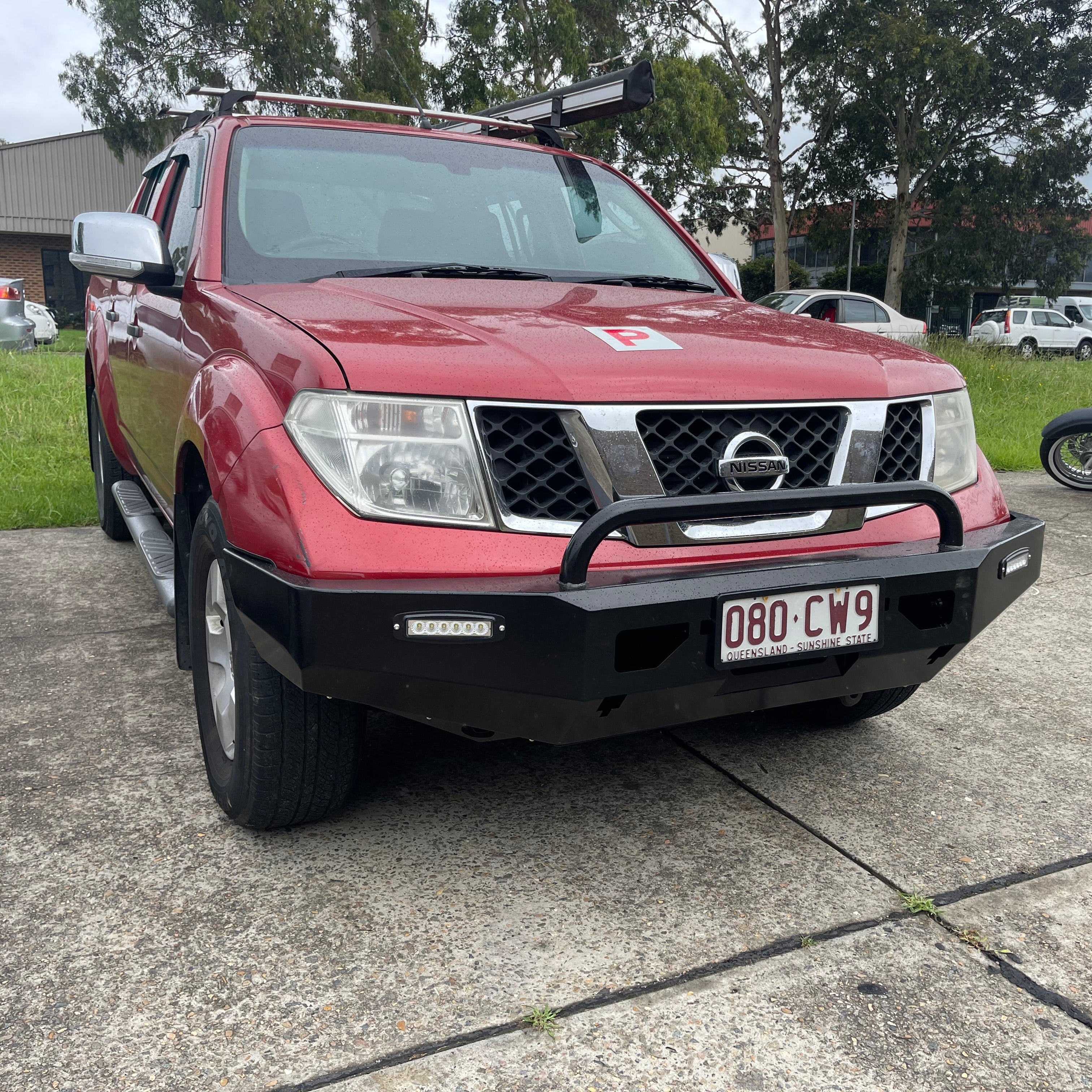A red Nissan Navara (D40 generation) pickup truck is parked on asphalt pavement, viewed from the front. It is equipped with a black, heavy-duty aftermarket steel bull bar/bumper that includes a single black tubular hoop and an integrated horizontal LED light bar on the driver's side. The truck has silver roof racks, silver side steps, a red 'P' plate (indicating a probationary driver) on the hood, and a black Australian license plate (080 CW9, QLD - Sunshine State) mounted on the bumper.