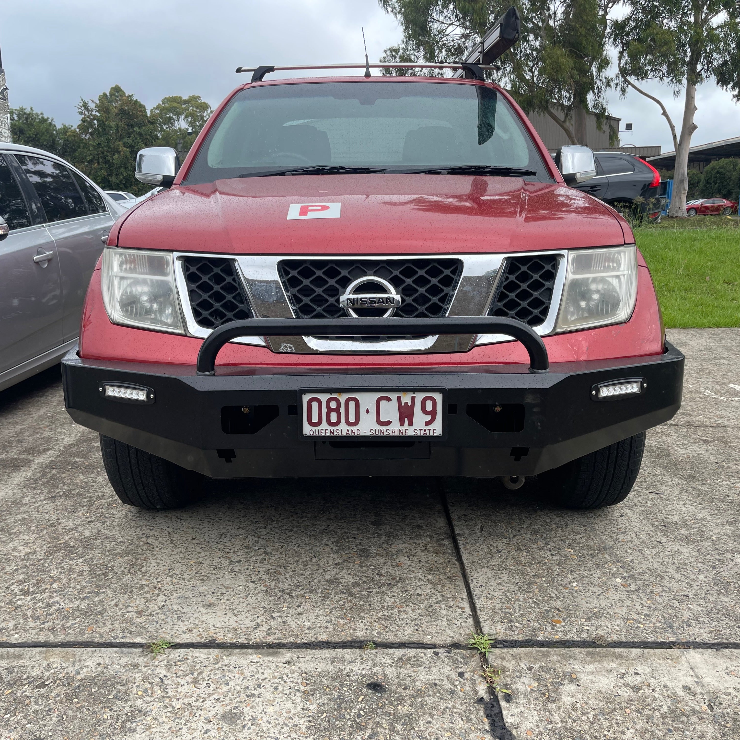 A head-on view of a red Nissan Navara (D40 generation) pickup truck parked on a concrete surface. The truck is fitted with a heavy-duty, angular black steel aftermarket bull bar/bumper. The bumper features a single tubular hoop, an integrated horizontal LED light bar on both the left and right sides, and an Australian license plate (080 CW9, QLD - Sunshine State) mounted centrally. The vehicle also has silver roof racks, silver mirror caps, and a red 'P' plate (indicating a probationary driver) placed on th