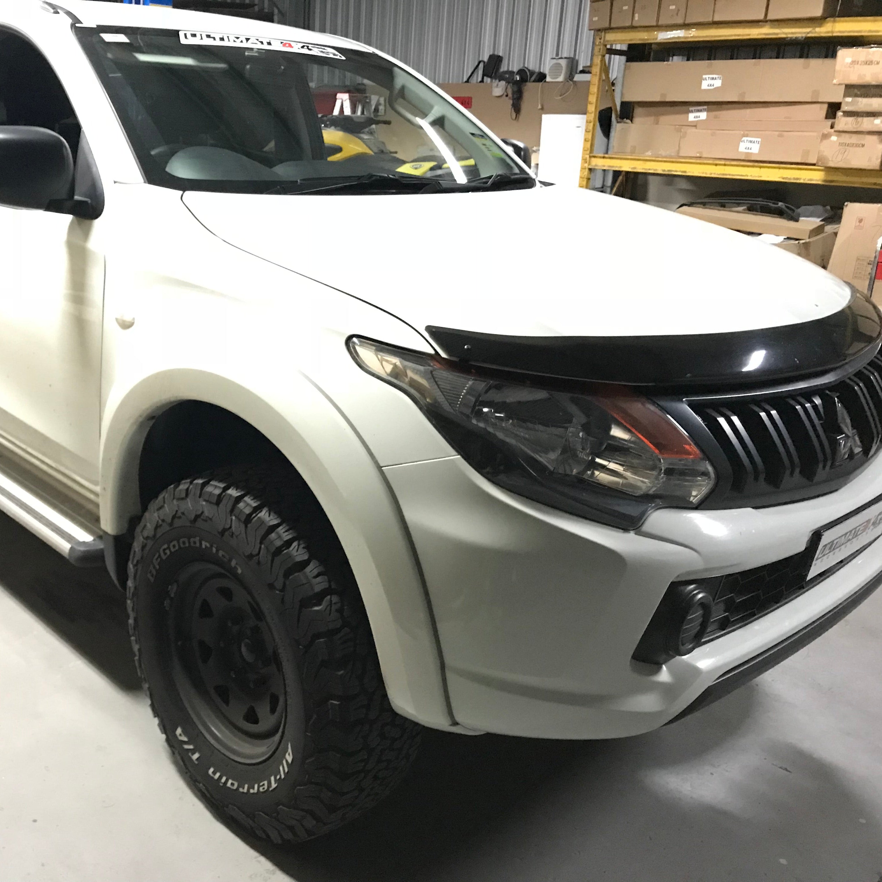 A white double-cab pickup truck (likely a Mitsubishi Triton/L200) shown from a front-three-quarter angle against a white background. The truck is fitted with several aftermarket accessories, including a black bonnet protector (bug shield) over the hood, a black grille, aggressive all-terrain tires on black steel wheels, and polished silver side steps/running boards.