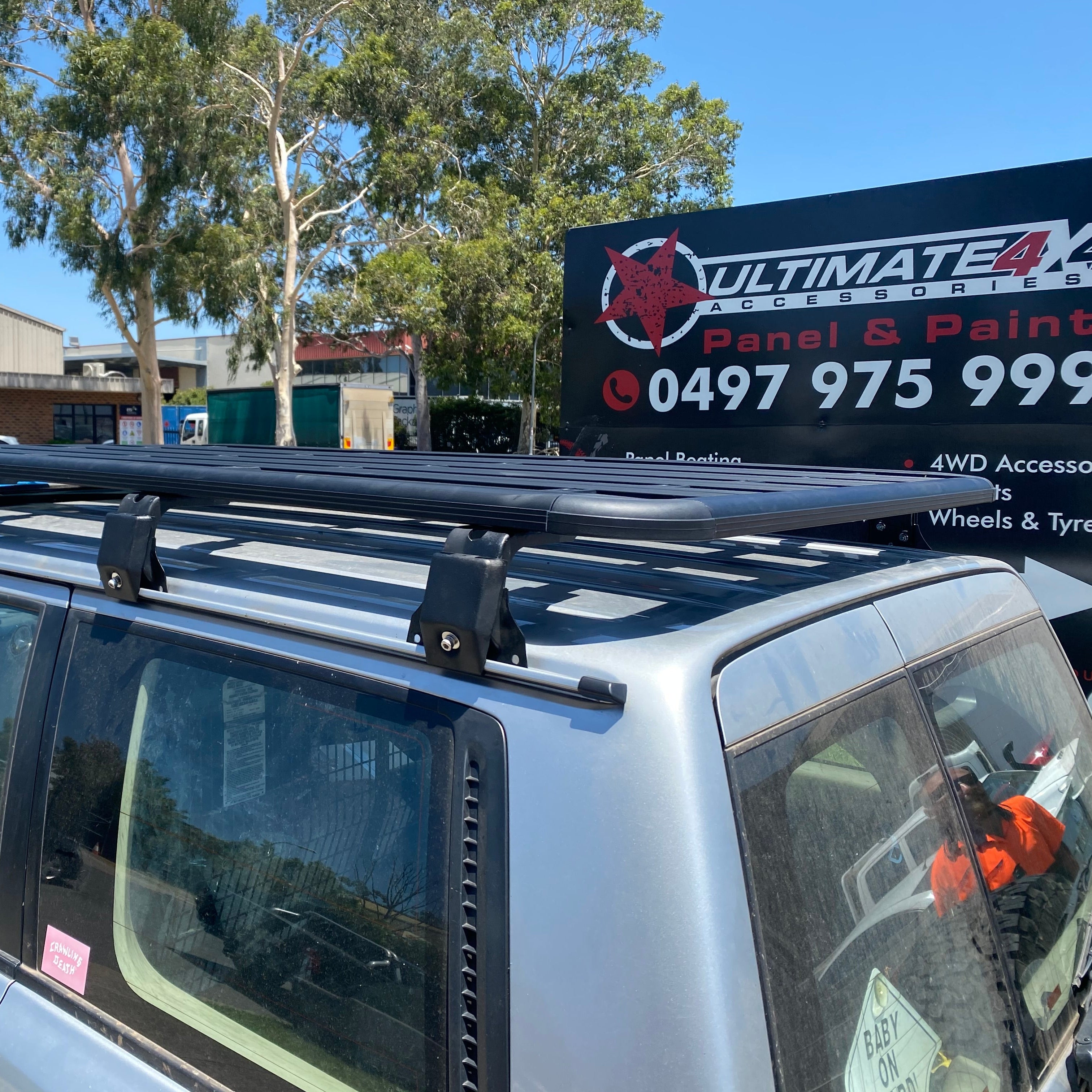 A close-up side view of the rear roof section of a silver SUV, highlighting a black platform-style roof rack mounted on the vehicle's rails. The mounting hardware and the slats of the rack are clearly visible. In the background, there is a large black sign for "ULTIMATE 4X4 ACCESSORIES Panel & Paint" with a phone number, and trees under a clear blue sky. A partial reflection of a person in an orange safety vest is visible on the rear window.