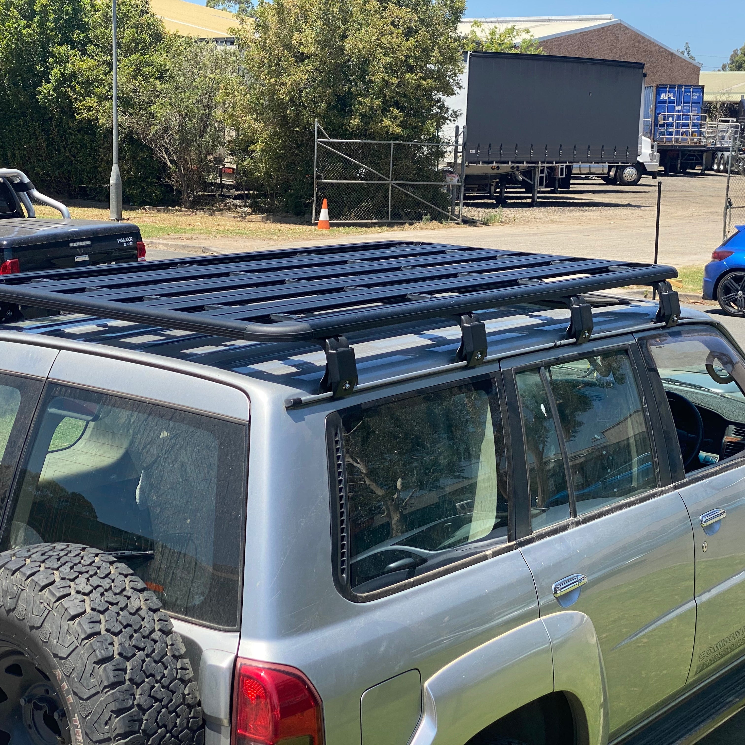 A side view of the rear section of a silver SUV, likely a Nissan Patrol, showing a large, rectangular black platform roof rack mounted on the vehicle's roof rails. An off-road tire is mounted on the rear door. The vehicle is parked outdoors, with trees, a fence, and several large trucks visible in the sunny background.