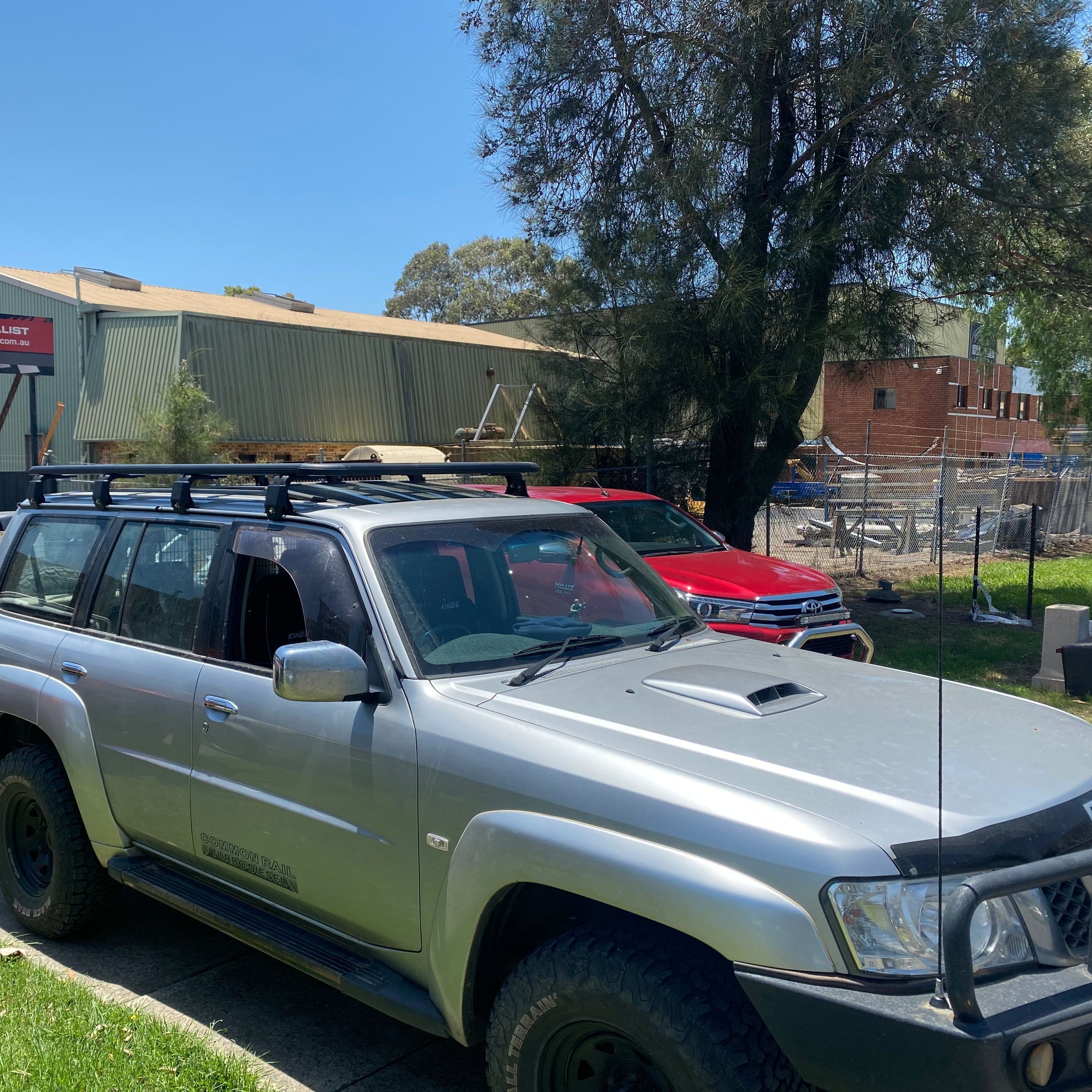 A close-up of a silver Nissan Patrol SUV, parked outdoors on a sunny day. The vehicle is equipped with a black roof rack, a snorkel, a black bull bar, and off-road tires. In the background, a red vehicle is partially visible, and behind them are a construction site with exposed brick and a light green industrial-style building.