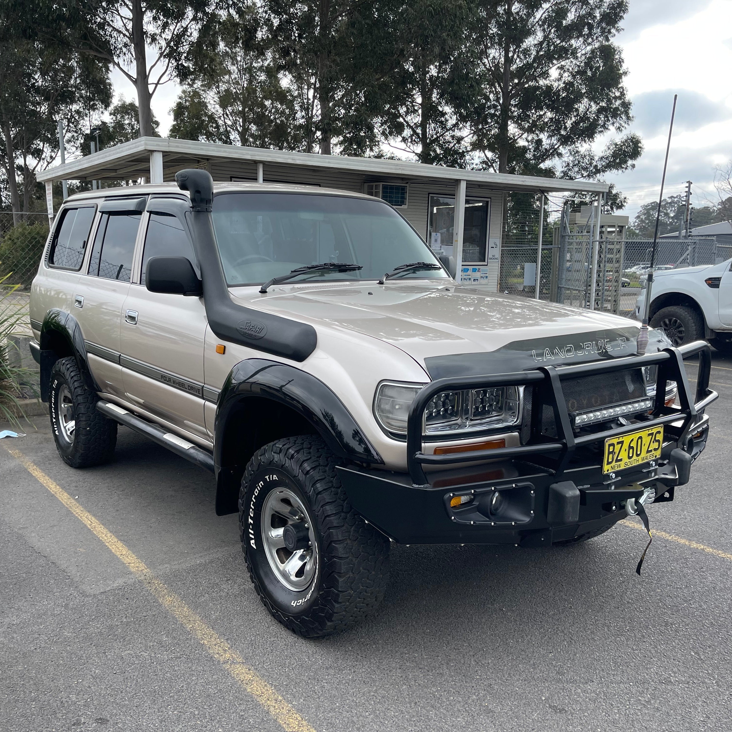 A highly modified champagne gold/beige Toyota Land Cruiser 80 series (FJ80/HZJ80) parked in a lot. The off-road vehicle features a heavy-duty black steel bull bar with an LED light bar, a black snorkel, black fender flares, and large BFGoodrich All-Terrain T/A tires on chrome wheels. It also has a hood protector, window visors, and New South Wales, Australia license plates reading "BZ 60 7S." The background includes trees and a light-colored outdoor building.