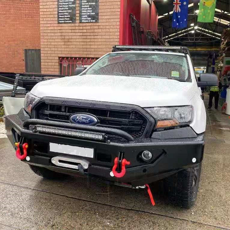 A white Ford Ranger pickup truck (likely 2015-2022 generation) is shown from a low front view, modified with a black, heavy-duty steel bull bar/bumper. The bumper features a central tubular hoop, a horizontal LED light bar mounted below the hoop, and a winch with a visible silver fairlead and rope. Two prominent red recovery shackles are attached to the bumper. The vehicle also has a roof rack with an additional black LED light bar.