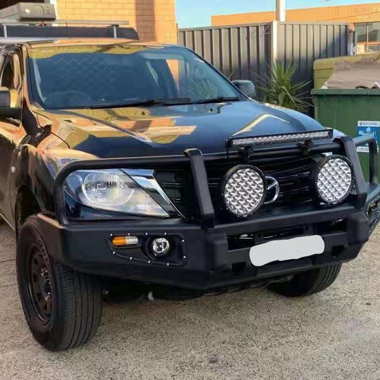
A black Mazda BT-50 pickup truck is parked outside, featuring an aftermarket, heavy-duty matte black steel bull bar. The bull bar has a triple-loop design protecting the grille and headlights. Two large, circular high-intensity driving lights with prominent, textured reflector faces are mounted prominently in front of the grille, attached to the middle bar of the bull bar. An LED light bar is also mounted just above the grille opening. 