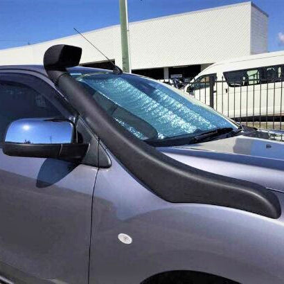 A close-up photograph of the front passenger side of a dark grey pickup truck (possibly a Ford Ranger or Mazda BT-50). The vehicle has a large black automotive snorkel installed, running from the fender up the A-pillar, with a square air ram head visible near the roofline. The truck also features a chrome side mirror and a reflective silver sun shade placed inside the windshield. In the blurred background, a white commercial van and a metal fence are visible.