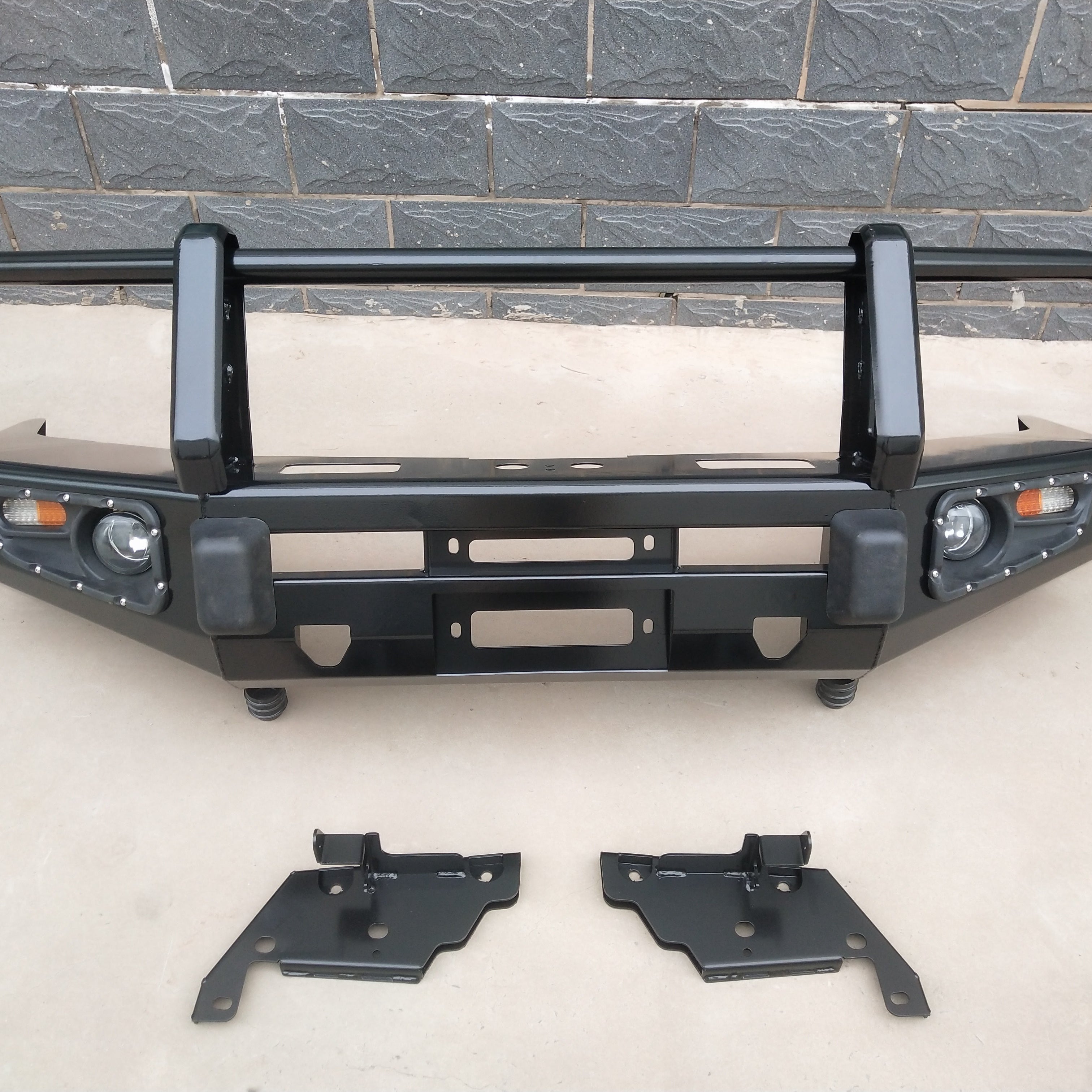 A heavy-duty, black metal off-road bull bar or bumper replacement is shown, designed for a truck or SUV. It features a triple-hoop design (one horizontal bar connecting two vertical uprights), integrated fog lights and turn signals on both sides, and a winch mounting plate in the center. Two separate black metal mounting brackets are lying on the ground in front of the bumper.