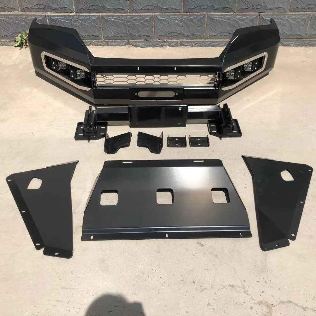 A black, heavy-duty aftermarket front bumper and skid plate kit for an off-road vehicle, displayed on a concrete surface in front of a dark gray cinder block wall. The main bumper features a sleek, angular design with a mesh grille insert, integrated square LED light housings accented with chrome or silver trim, and a winch fairlead opening. Below the bumper are various mounting brackets and a winch plate. At the bottom, three large, separate black metal plates form the skid plate assembly.