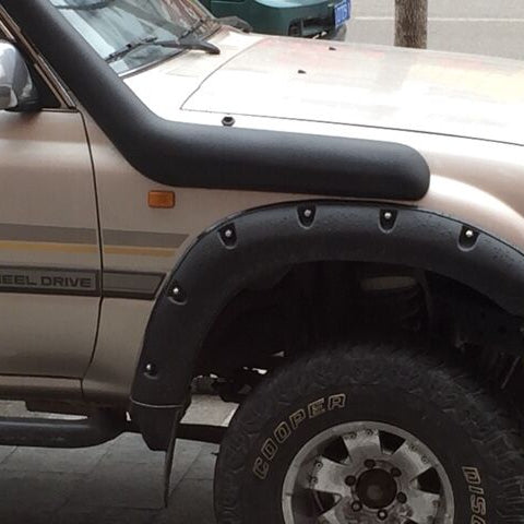 A close-up side view of the front fender and bonnet of a tan/beige older model Toyota Land Cruiser (likely an 80 series). The truck is modified with a large, curved black plastic snorkel installed across the driver's side fender and a wide, black plastic fender flare with exposed silver bolt heads over the wheel arch. The tire is a Cooper brand all-terrain or mud-terrain model on a silver alloy rim.