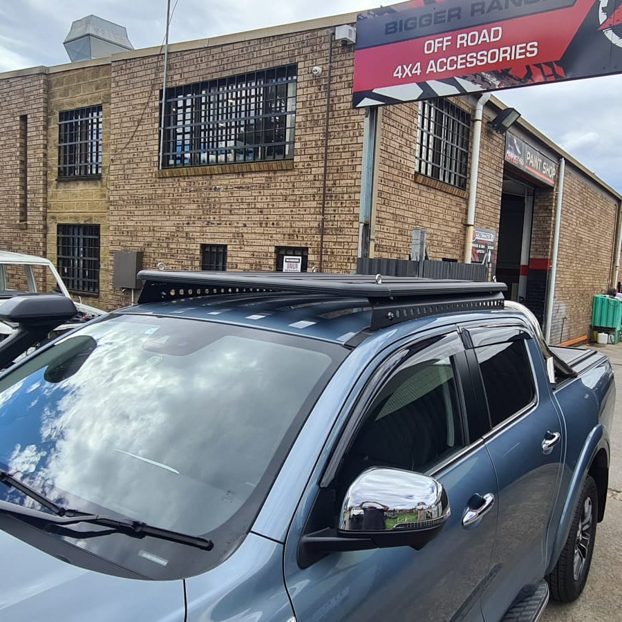 A medium-angle photo focusing on the roof of a metallic blue pickup truck, likely a GWM Cannon or Nissan Navara, parked in front of a brick building. The truck is fitted with a low-profile black platform roof rack on the cab. The sky with white clouds is reflected on the windshield and the chrome side mirror. A red and white sign for "OFF ROAD 4X4 ACCESSORIES" is visible above the truck.