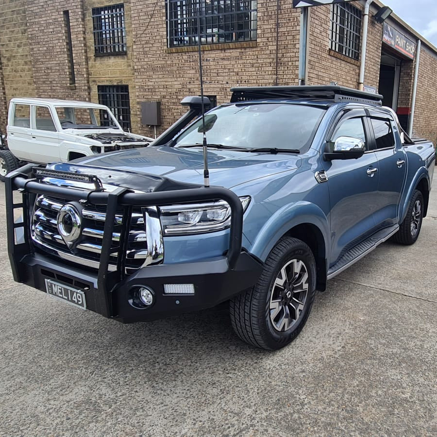 A slightly low-angle photo of a heavily modified, metallic blue pickup truck, likely a GWM Cannon, parked in an industrial area. The truck is fitted with a large black steel bull bar on the front, a snorkel, a platform roof rack on the cab, and a long antenna. A white classic 4x4, possibly a Toyota Land Cruiser, is parked to the left behind the blue truck. The background is a brick building labeled 'Paint Shop'.