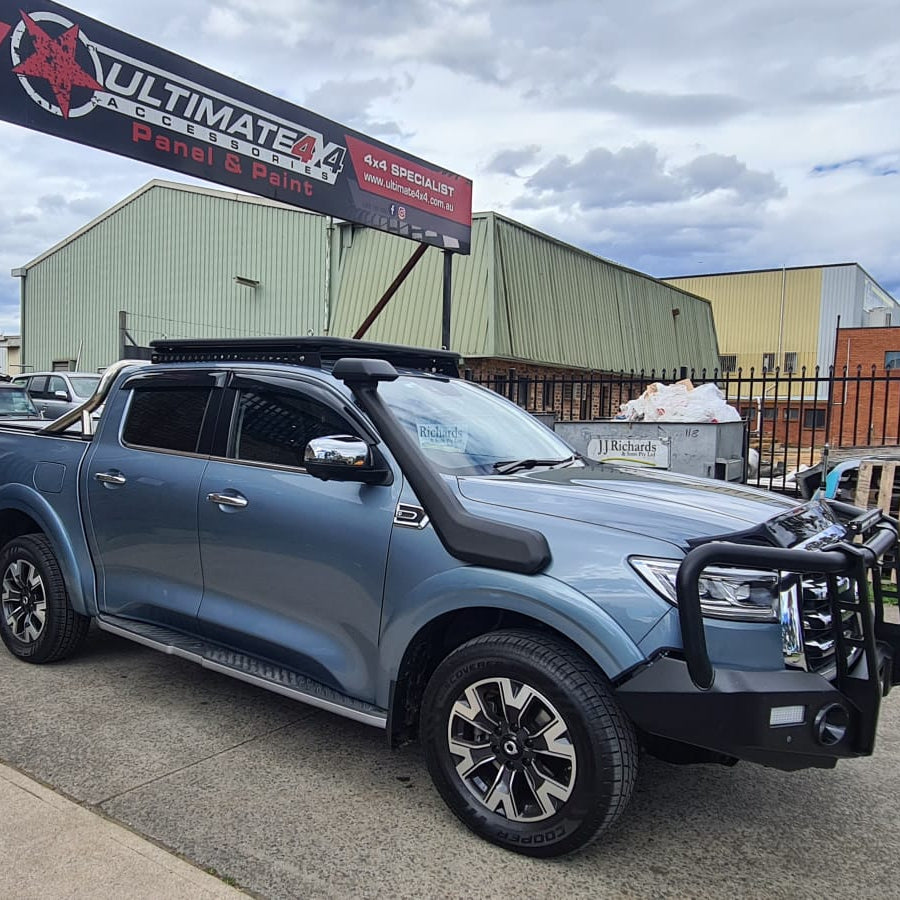 A photo of a metallic blue pickup truck, likely a GWM Cannon, parked outside an automotive shop specializing in 4x4 accessories. The truck is heavily accessorized with a large black steel bull bar, a prominent black snorkel on the passenger side, a platform roof rack on the cab, and side steps. A red and black banner reading "ULTIMATE 4X4 ACCESSORIES Panel & Paint" hangs above the truck. The background shows industrial buildings under an overcast sky.