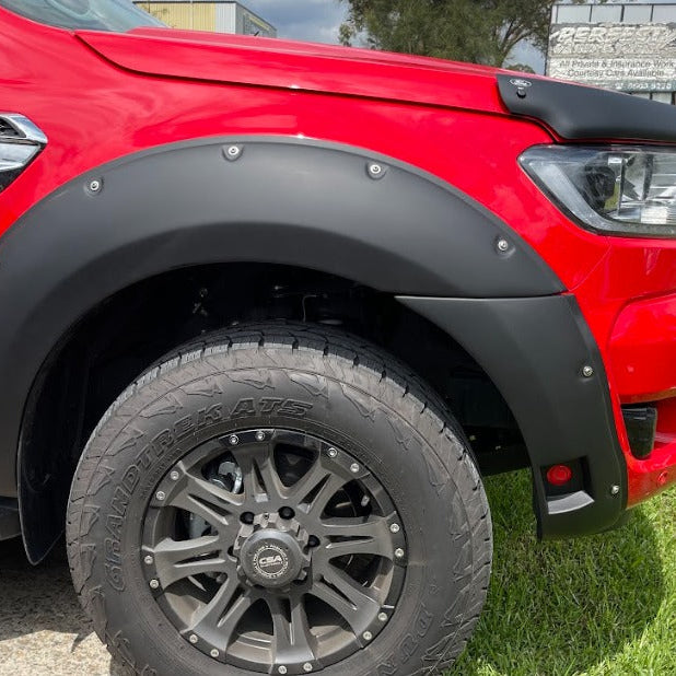 A close-up view of the front wheel arch of a bright red pickup truck (likely a Ford Ranger) fitted with a matte black bolt-on style fender flare. The flare has visible silver rivets along the top edge. The truck has a black multi-spoke wheel with an off-road tire (Dunlop Grandtrek AT3). A black bonnet/hood protector is also visible near the windshield.