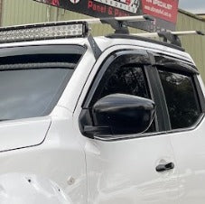A close-up image of the front door and cab of a white pickup truck, showing several installed accessories. These accessories include a large LED light bar mounted above the windshield, black and silver roof rack cross bars, dark plastic window visors/rain guards, and a black side mirror. A red and white sign is visible at the very top.