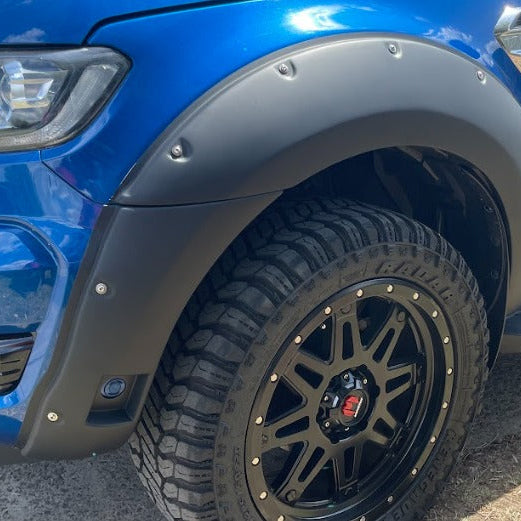 A tight close-up view of the front wheel arch of a vibrant blue pickup truck. The truck is equipped with a wide, matte black bolt-on style fender flare that extends onto the bumper. The flare has visible silver rivets along the top and bottom edge. The wheel is a black multi-spoke off-road rim fitted with a large, aggressive mud-terrain tire.