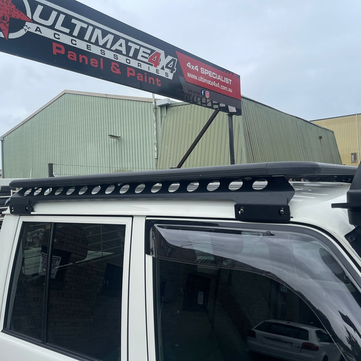 A tight side view focusing on a black, flat platform roof rack mounted on the white roof of a vehicle. The image clearly shows the slotted side rail design and the black mounting brackets securing the rack to the vehicle's gutter. In the blurred background, a sign for "ULTIMATE 4X4 ACCESSORIES Panel & Paint" is visible above industrial buildings.