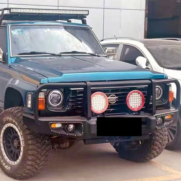 A close-up view of the front end of a modified, dark teal or blue-green Nissan Patrol Y60/GQ SUV. The vehicle is equipped with a heavy-duty, matte black steel bull bar featuring a triple hoop design, two large circular LED driving lights with red accents, and aftermarket LED projector headlights. A long LED light bar is visible mounted on a roof rack above the windshield. The foreground shows aggressive, chunky mud-terrain tires on black aftermarket wheels.