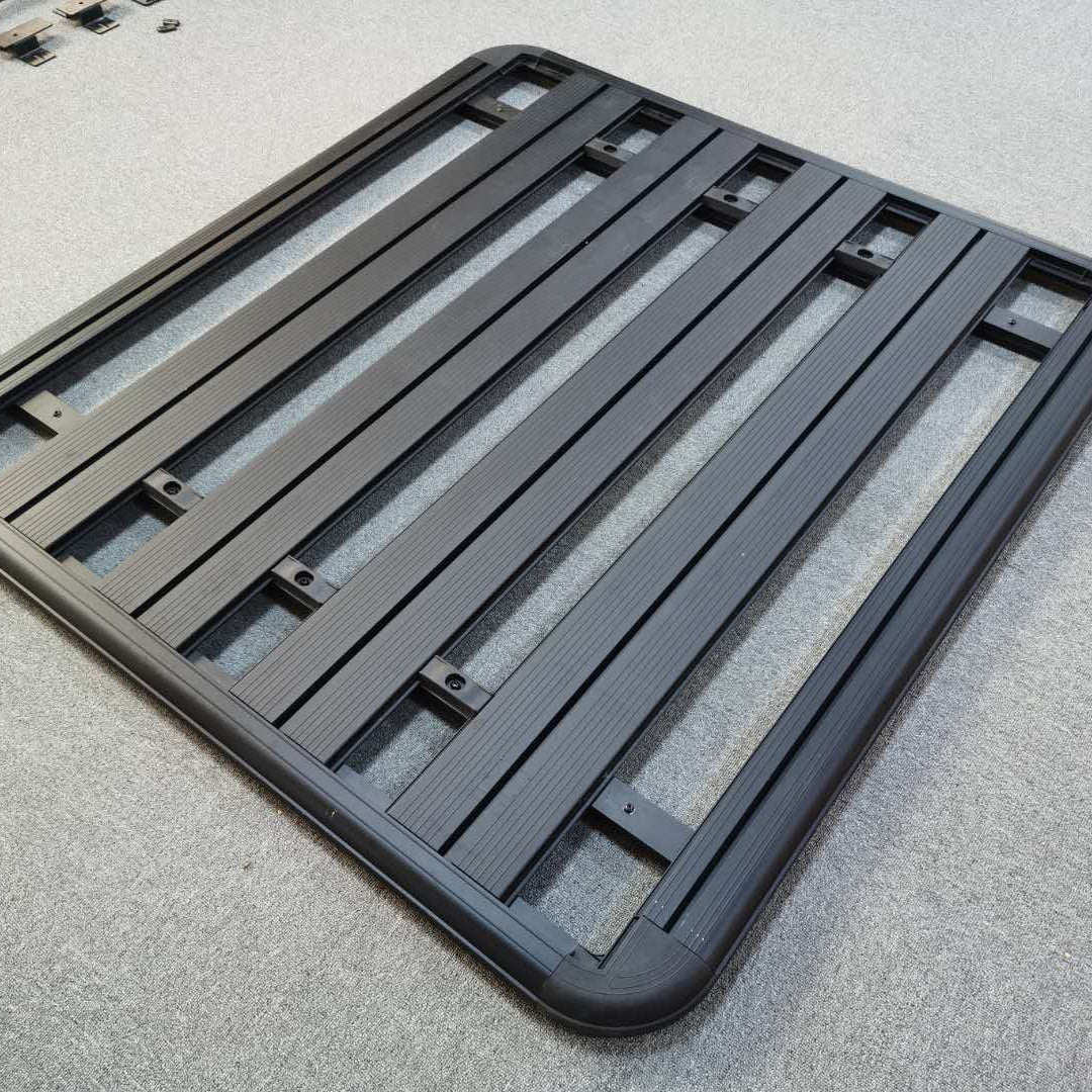A slightly angled view of a rectangular, black, flat-platform style vehicle roof rack. The rack is made of thick aluminum or steel and features multiple parallel slats/crossbars set into a sturdy, rounded outer frame. It is resting on a light gray carpeted surface.